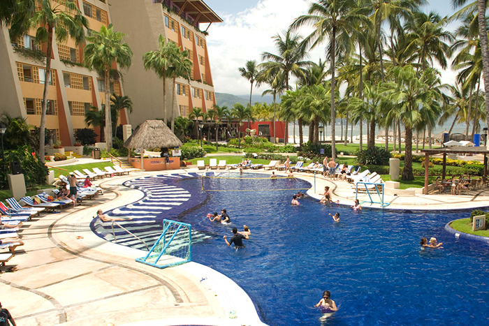 Fotografía que muestra la alberca de un hotel donde se aprecia el hotel del costado izquierdo y del derecho palmeras